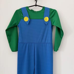 May include: A long-sleeved jumpsuit featuring a green shirt top integrated with blue overalls. The overalls have two wide shoulder straps, each adorned with a circular yellow button detail at the front, creating a distinct two-tone color block design.