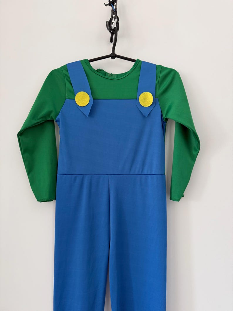 May include: A long-sleeved jumpsuit featuring a green shirt top integrated with blue overalls. The overalls have two wide shoulder straps, each adorned with a circular yellow button detail at the front, creating a distinct two-tone color block design.