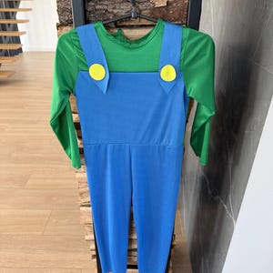 May include: A long-sleeved jumpsuit featuring a green shirt base with an attached blue overall overlay. The blue overalls have two wide shoulder straps, each accented with a large, circular yellow button at the front, creating a distinct two-tone look.
