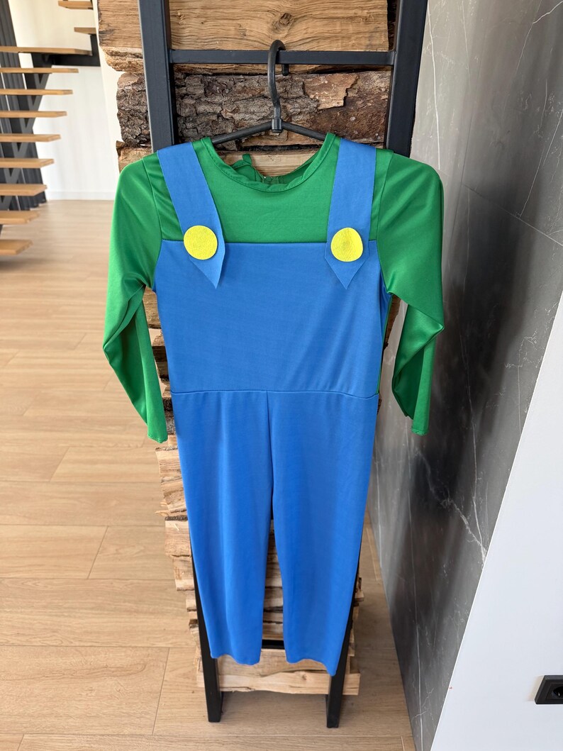 May include: A long-sleeved jumpsuit featuring a green shirt base with an attached blue overall overlay. The blue overalls have two wide shoulder straps, each accented with a large, circular yellow button at the front, creating a distinct two-tone look.