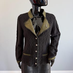 May include: A black jacket with a textured design, olive green collar and trim, and silver buttons. A small silver brooch is pinned on the lapel. The jacket is displayed on a black mannequin.