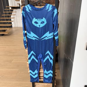 May include: A blue jumpsuit with a diamond pattern and light blue accents. The jumpsuit features a cat-like mask design on the chest and lightning bolt shapes. The long-sleeved outfit is hanging on a black rack.
