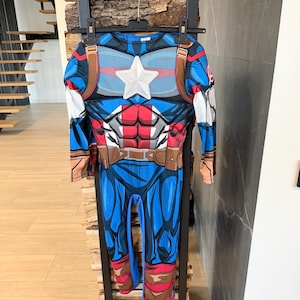 May include: A long-sleeved Captain America costume jumpsuit featuring a blue, red, and white color scheme. The chest displays a prominent white star, printed abdominal muscle details, and a brown utility belt graphic around the waist area.