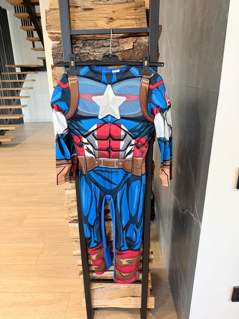 May include: A long-sleeved Captain America costume jumpsuit featuring a blue, red, and white color scheme. The chest displays a prominent white star, printed abdominal muscle details, and a brown utility belt graphic around the waist area.