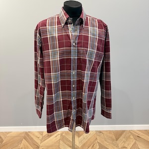 May include: A burgundy, white, blue, and tan plaid long-sleeved shirt, buttoned up and displayed on a mannequin. The shirt has a classic collar and a single chest pocket, typical of a casual button-down.