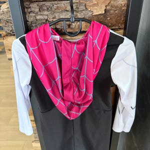 May include: A black and white superhero costume with a pink hood featuring a blue spiderweb design. The long-sleeved costume is hanging on a black hanger against a wooden background. The costume is designed for children.