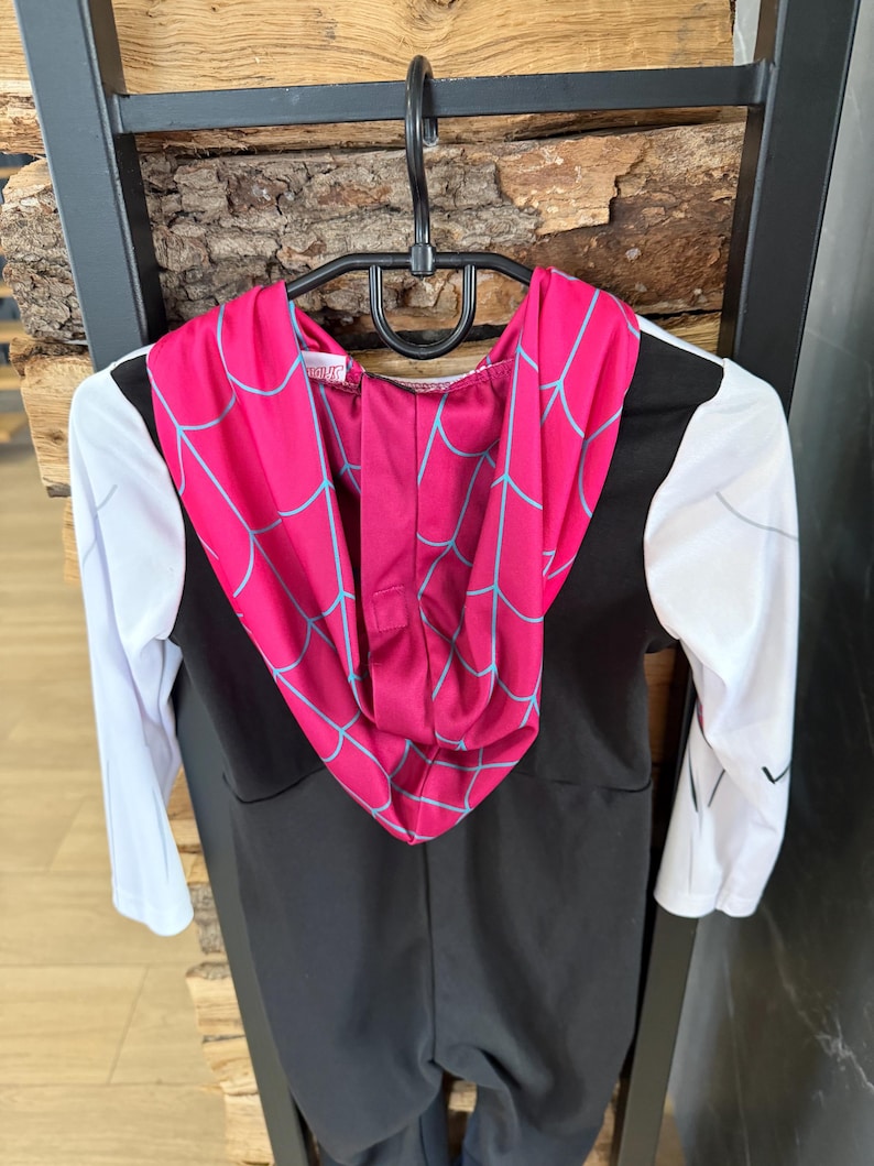 May include: A black and white superhero costume with a pink hood featuring a blue spiderweb design. The long-sleeved costume is hanging on a black hanger against a wooden background. The costume is designed for children.