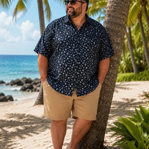 May include: A short-sleeved button-down shirt in navy blue with a white repeating pattern of small tropical icons, paired with solid tan khaki shorts and brown flip-flop sandals. The shirt features a classic collar and a full front button placket.