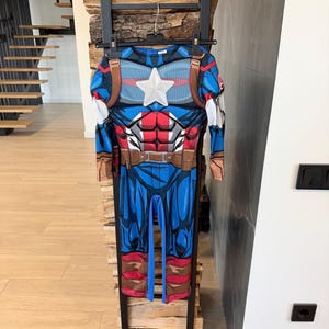 May include: A full-body Captain America costume jumpsuit featuring a blue, red, and white color scheme with a printed muscular torso design, a prominent white star on the chest, brown utility belt details, and long sleeves with attached gauntlets.