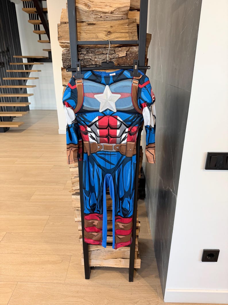 May include: A full-body Captain America costume jumpsuit featuring a blue, red, and white color scheme with a printed muscular torso design, a prominent white star on the chest, brown utility belt details, and long sleeves with attached gauntlets.
