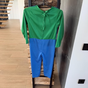 May include: A long-sleeved jumpsuit featuring a vibrant green top section and a bright blue bottom section. The garment includes a round neckline with a tie closure at the back of the neck, and it is displayed on a black plastic hanger.