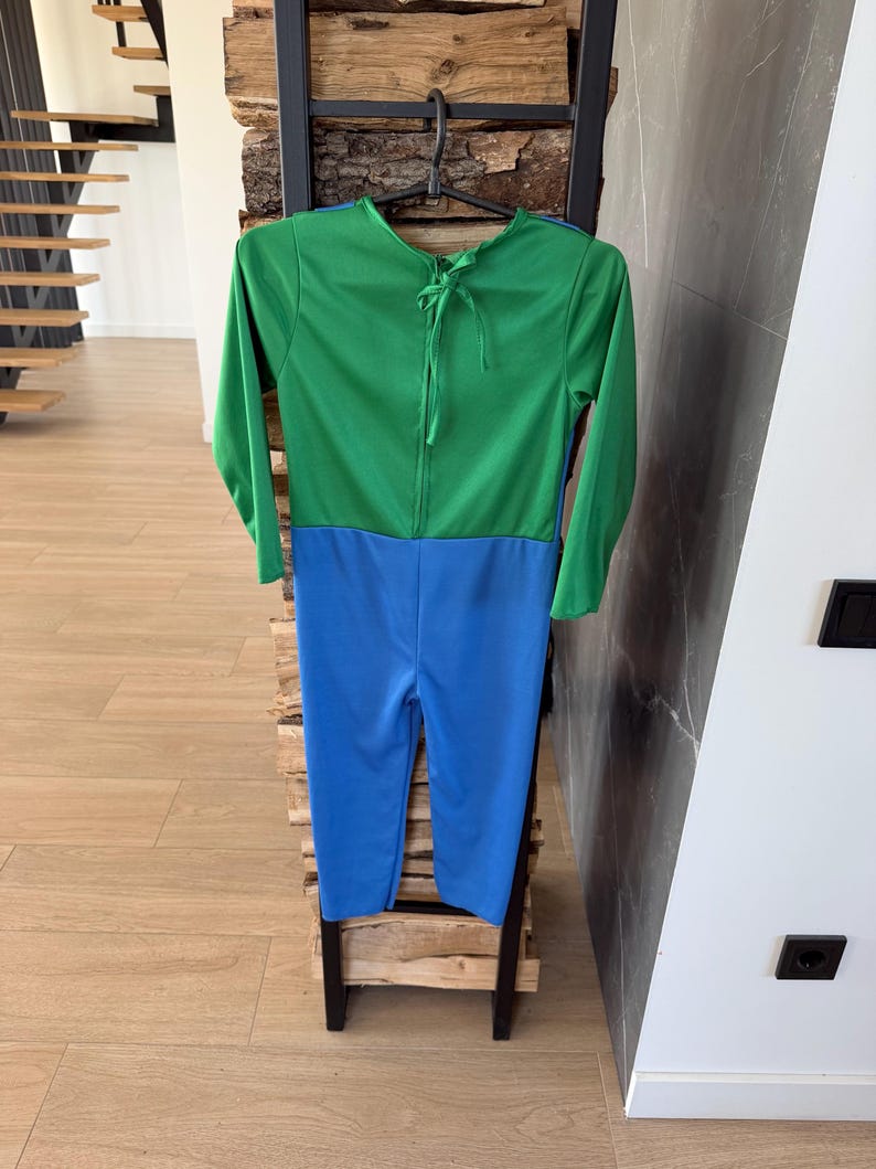 May include: A long-sleeved jumpsuit featuring a vibrant green top section and a bright blue bottom section. The garment includes a round neckline with a tie closure at the back of the neck, and it is displayed on a black plastic hanger.