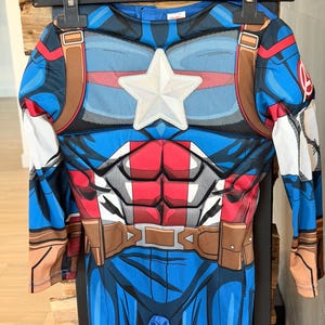 May include: A long-sleeve Captain America costume jumpsuit featuring a blue, red, and white color scheme. The chest displays a prominent white star emblem, printed abdominal muscle details, and a brown utility belt graphic around the waist.