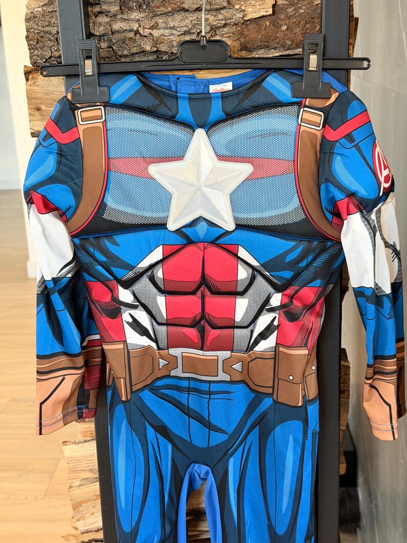 May include: A long-sleeve Captain America costume jumpsuit featuring a blue, red, and white color scheme. The chest displays a prominent white star emblem, printed abdominal muscle details, and a brown utility belt graphic around the waist.