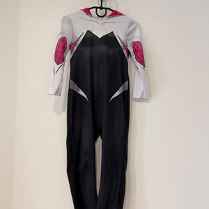 May include: A Spider-Gwen costume, a white and black jumpsuit with pink elbow patches. The costume has a red-lined hood and is hanging on a black hanger. The design is based on the character's distinctive appearance, hanging against a white wall.