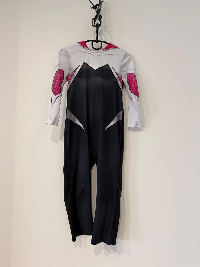 May include: A Spider-Gwen costume, a white and black jumpsuit with pink elbow patches. The costume has a red-lined hood and is hanging on a black hanger. The design is based on the character's distinctive appearance, hanging against a white wall.