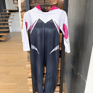 May include: A Spider-Gwen costume in white, black, and pink, hanging on a black hanger. The long-sleeved jumpsuit has a white upper section with black and pink details, and a black lower section. The costume is displayed against a wooden and black metal backdrop.