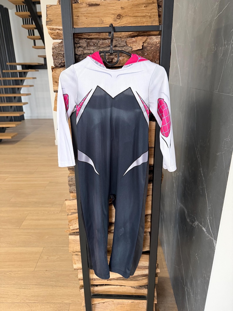 May include: A Spider-Gwen costume in white, black, and pink, hanging on a black hanger. The long-sleeved jumpsuit has a white upper section with black and pink details, and a black lower section. The costume is displayed against a wooden and black metal backdrop.