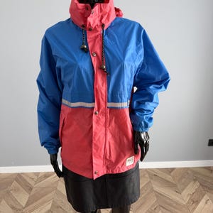Könnte beinhalten: Blau-korallenfarbene Jacke mit hohem Kragen und reflektierendem Streifen. Die Jacke hat eine Kapuze mit Kordelzug und eine Knopfleiste. Die Jacke wird auf einer Schaufensterpuppe präsentiert.