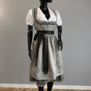 Op de afbeelding: Een dirndl-jurk met een witte blouse met pofmouwen, een grijs tweed lijfje met zwarte en parelmoeren afwerking, en een lichtgrijze rok met een zwarte satijnen linttaille. De rok heeft een subtiele glans en een donkergrijze zoomafwerking.