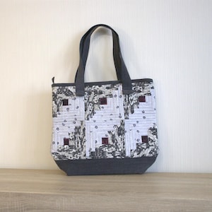 May include: A quilted tote bag with a gray base and handles. The body of the bag features a patchwork design in shades of gray, black, and white, with small burgundy squares. The bag is sitting on a wooden surface.