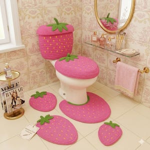 Puede incluir: Un juego de baño rosa tejido a ganchillo con temática de fresa, que incluye una funda para el asiento del inodoro, una funda para la tapa y alfombras. El juego presenta hojas verdes y detalles de semillas amarillas.