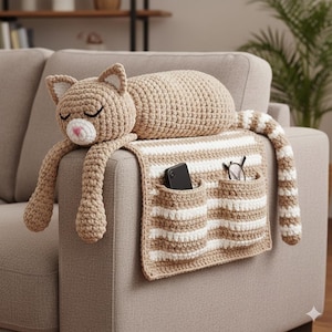 Puede incluir: Un cojín con forma de gato y un organizador de reposabrazos de ganchillo a rayas beige y blancas. El gato tiene los ojos cerrados, una nariz rosa y brazos extendidos. El organizador tiene dos bolsillos con un teléfono y gafas.