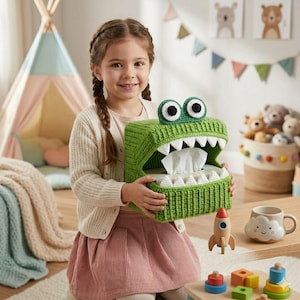 May include: A green crocheted tissue box cover shaped like a crocodile with white teeth and large, round eyes. The tissue box is held by a person in a pink skirt and cream cardigan. The tissue box is filled with white tissues.