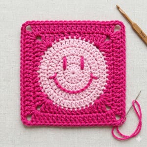 May include: A handmade, square crochet piece featuring a smiling face design. The square is a vibrant fuchsia pink, with a light pink smiley face in the center. A crochet hook and needle with pink thread are also visible.