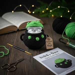 Puede incluir: Una funda para AirPods de ganchillo negro con forma de caldero con detalles verdes y una tapa verde, que contiene AirPods blancos. Una etiqueta dice "Handmade with Magic". Un patrón de ganchillo "Witch's Brew" es visible.
