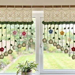 May include: Handmade crochet window curtains with a floral design. The curtains feature a cream-colored lace top, green trim, and hanging strands with colorful flowers in red, blue, yellow, and pink. The curtains are hung on a wooden rod.