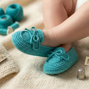 May include: A pair of turquoise crochet baby loafers with bow details. The shoes are on a cream-colored blanket next to a matching knitted hat and spools of turquoise thread. The label reads "BABY LOAFERS - 0-6M".