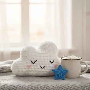 May include: A white crochet cloud-shaped pillow with a smiling face, a blue star, and a mug of hot chocolate with marshmallows. The items are on a gray blanket near a window.