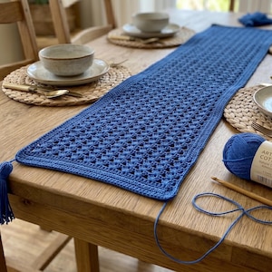 Może przedstawiać: Niebieski, szydełkowy bieżnik z frędzlami, ustawiony na drewnianym stole. Kłębek przędzy Artisan Cotton, szydełko i ołówek leżą obok bieżnika. Stół jest nakryty miskami, talerzami i podkładkami.