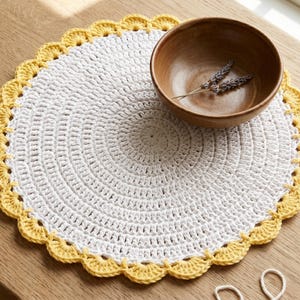 May include: A handmade, white crochet placemat with a scalloped yellow border. A wooden bowl with dried lavender sits on the placemat, displayed on a wooden table. Crochet hooks and yarn are also visible.