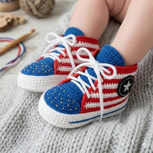 Peut inclure: Une paire de baskets pour bébé crochetées à la main avec un motif rayé rouge et blanc, des bouts bleus avec de petits clous dorés, des lacets blancs et un écusson circulaire noir sur le côté avec une étoile blanche, portées aux pieds.