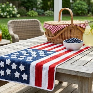 May include: A wooden picnic table is set for a summer gathering. A crocheted table runner in the style of the American flag is laid across the table. A wicker picnic basket, two pitchers of lemonade, and a bowl of blueberries complete the scene.