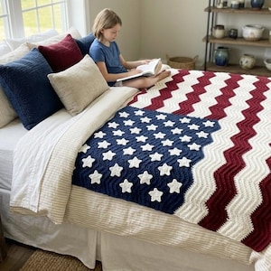 Könnte beinhalten: Eine gehäkelte Decke mit einem Zickzackmuster in Rot, Weiß und Blau sowie weißen Sternapplikationen in einem rechteckigen Feld. Die Decke liegt auf einem Bett mit weißer Bettwäsche und wird von mehreren dekorativen Kissen in Unifarben ergänzt.