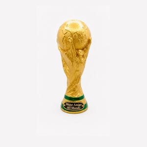 Könnte beinhalten: Eine goldfarbene Trophäe, die einen von zwei Figuren getragenen Globus darstellt. Der Sockel ist mit einem grünen Band und einer schwarzen Plakette versehen, die den Text "Mejor Amigo del Mundo" in goldener Schrift zeigt. Die Trophäe hat ein metallisches Finish.