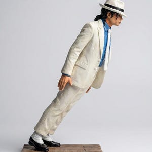 Può includere: Una statuetta di un artista inclinato in avanti con un angolo estremo, che indossa un abito bianco, una camicia blu, una cravatta bianca e un cappello fedora bianco con una fascia nera. La figura ha capelli scuri ed è su una base di legno.
