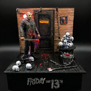 Puede incluir: Un diorama de Friday the 13th con una figura de Jason Voorhees con máscara de hockey sosteniendo un machete. La escena incluye una pared de madera con un letrero de Camp Crystal Lake, calaveras, cadenas, un cubo de sangre y un hacha.