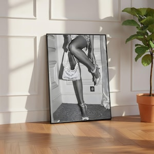May include: Black and white framed print featuring a person wearing a sequined outfit, holding a sequined handbag, and wearing high heels with rhinestone embellishments. The print is leaning against a white wall and a wooden floor.