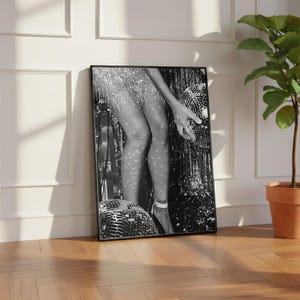 May include: Black and white framed print featuring a person in a sparkling dress holding a disco ball. The image has a vintage aesthetic, with light reflecting off the disco ball and the person's legs. The print is leaning against a white wall.