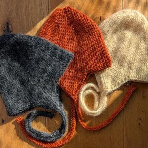 May include: Three knitted baby bonnets in shades of charcoal, burnt orange, and cream. Each bonnet features a rounded crown and ties with long, braided cords. The bonnets are displayed on a wooden surface.