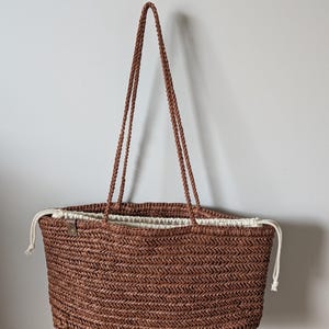 Puede incluir: Un bolso tote marrón tejido con patrón de espiga, dos asas largas trenzadas y un cierre de cordón de tela color crema en la parte superior. Una pequeña etiqueta rectangular está sujeta en el lado superior izquierdo del exterior.