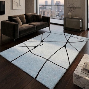 May include: A rectangular area rug with a light blue background and a black geometric design. The rug is placed on a dark wood floor in a modern living room setting. A brown sofa and a city view are visible.