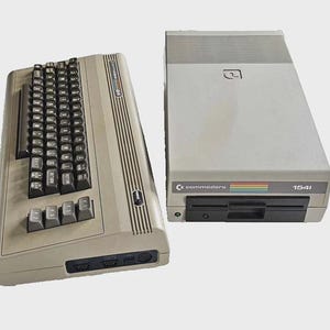 May include: A vintage Commodore 64 computer and a 1541 disk drive. The computer has a keyboard with black keys and a beige case. The disk drive is gray with a black disk drive slot and the Commodore logo.