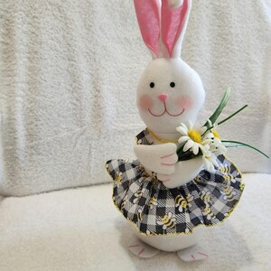 May include: A white plush bunny figurine with pink-lined ears, rosy cheeks, and a pink nose. The bunny wears a black and white checkered dress with yellow bees and holds a bouquet of white daisies. A charming Easter decoration.
