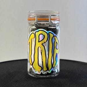 Op de afbeelding: Een doorzichtige glazen pot met een metalen beugelsluiting en een oranje rubberen afdichting. Op de voorkant van de pot zit een holografische sticker met het woord TRIP in een geel, druipend, vloeistofachtig lettertype op een achtergrond.