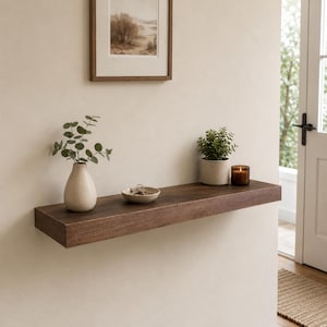 May include: A dark brown wooden floating shelf mounted on a beige wall. On the shelf sits a beige ceramic vase with green eucalyptus stems, a small beige bowl containing metal keys, a green potted plant, and a brown glass candle holder.